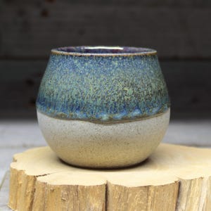 May include: A small, round ceramic cup with a blue and green glaze on the top half and a white glaze on the bottom half. The cup is sitting on a wooden surface.