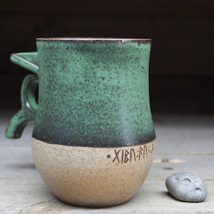 May include: A green and beige ceramic mug with a handle and a design on the bottom that reads "XIBN-MAGA".