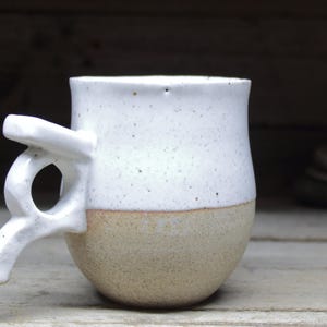 May include: A handmade ceramic mug with a unique handle design. The top half is white with speckled details, while the bottom half is a natural tan color. The handle has an artistic, open design. A rustic, handcrafted piece.