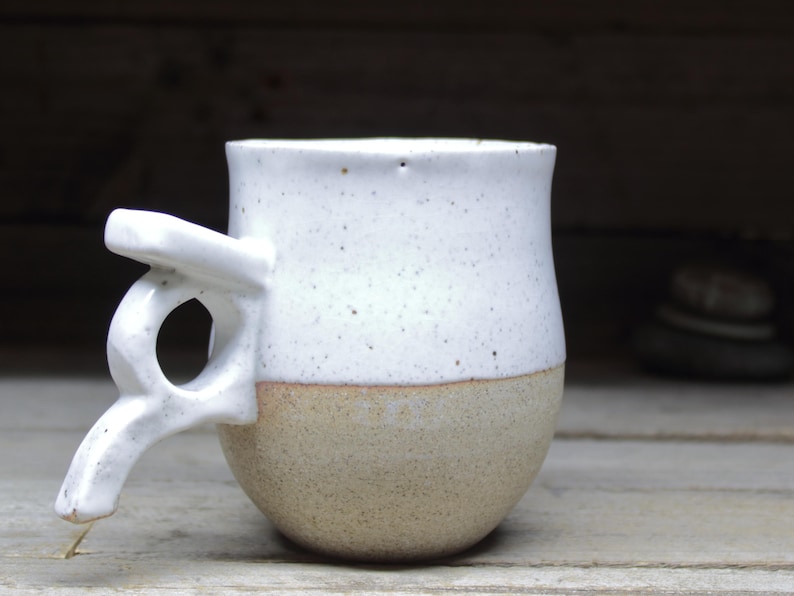 May include: A handmade ceramic mug with a unique handle design. The top half is white with speckled details, while the bottom half is a natural tan color. The handle has an artistic, open design. A rustic, handcrafted piece.