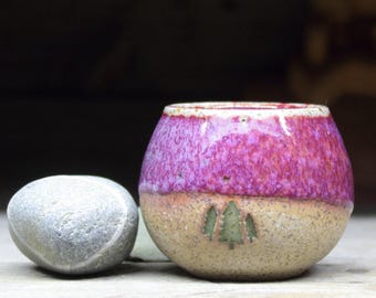 May include: A small, round ceramic bowl with a pink and brown glaze. The bowl has a design of three green trees on the bottom.