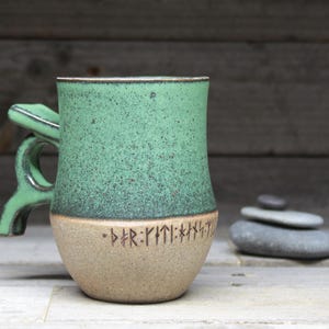 May include: A green ceramic mug with a unique handle and a beige bottom with runes inscribed on it.