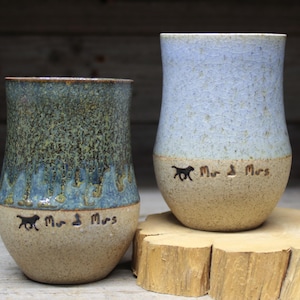 Może przedstawiać: Dwa ceramiczne kubki z jasnoniebieską i brązową glazurą. Kubki są ozdobione napisem "Mr. & Mrs." i odciskiem łapy.