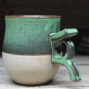 May include: A handmade ceramic mug with a unique handle design. The mug features a two-tone color scheme, with a speckled green upper half and a beige lower half. The handle is a matching green color.