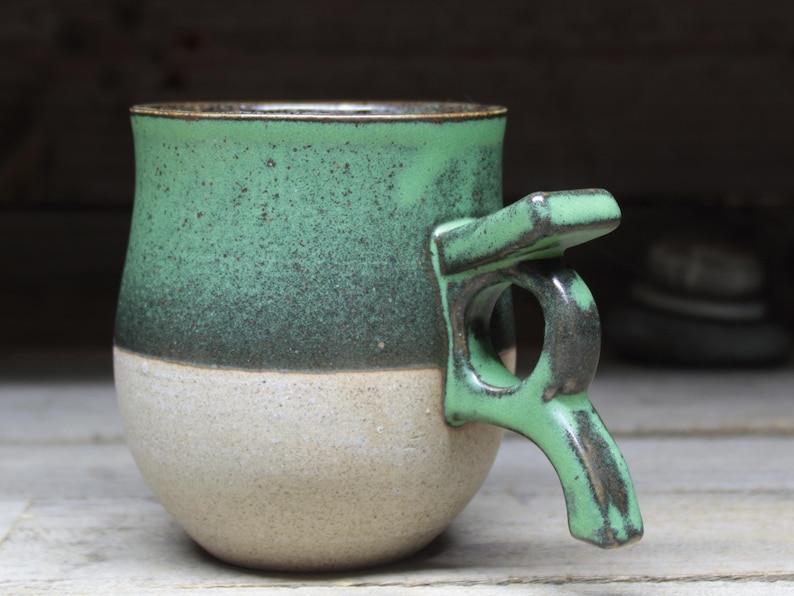 May include: A handmade ceramic mug with a unique handle design. The mug features a two-tone color scheme, with a speckled green upper half and a beige lower half. The handle is a matching green color.