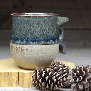 May include: A handcrafted ceramic mug with a blue and beige speckled glaze. The mug features a unique handle design and etched runes near the base. The mug is displayed on a wooden slice with pine cones.