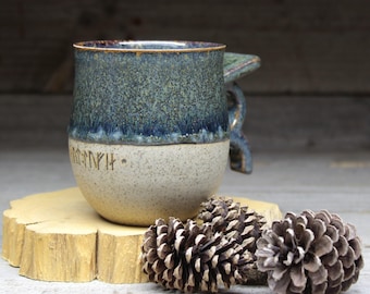 May include: A handcrafted ceramic mug with a blue and beige speckled glaze. The mug features a unique handle design and etched runes near the base. The mug is displayed on a wooden slice with pine cones.