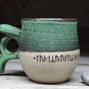 May include: A handmade ceramic mug with a unique green handle and a two-tone design. The top half is green, the bottom is beige, and it features etched runes. A small, gray stone with holes sits beside the mug.