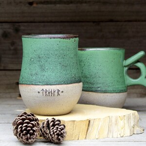 May include: Two ceramic mugs with green glaze and a white base. One mug has a handle and the other has a rune inscription that reads "TRIHKR".