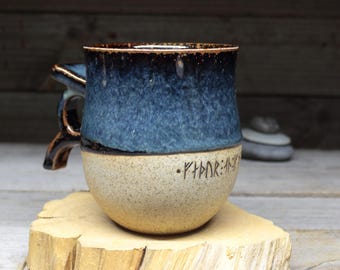 May include: A handmade ceramic mug with a blue and tan glaze. The top half is a deep blue, transitioning to tan at the base. The mug features a handle and etched runes. The mug is sitting on a wooden surface.