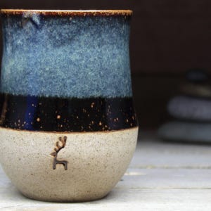 Könnte beinhalten: Eine Keramiktasse mit blauer, schwarzer und weißer Glasur. Die Tasse hat eine kleine Hirschsilhouette auf der Vorderseite.
