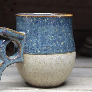 May include: Handmade ceramic mug with a unique handle design. The upper portion is a speckled blue, while the lower half is a speckled beige. The rim and handle have a brown glaze. A rustic, handcrafted piece for enjoying beverages.