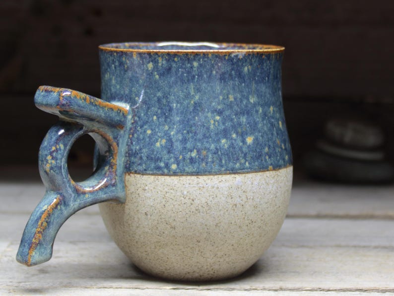 May include: Handmade ceramic mug with a unique handle design. The upper portion is a speckled blue, while the lower half is a speckled beige. The rim and handle have a brown glaze. A rustic, handcrafted piece for enjoying beverages.