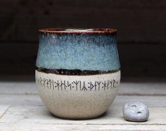 May include: A handmade ceramic tumbler with a blue and brown glazed top, a black band, and a beige base. The base features dark blue runic symbols. A small, gray stone sits beside the tumbler.