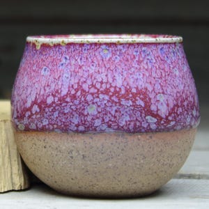 May include: A small, round ceramic pot with a speckled, reddish-purple glaze on the top half and a speckled, brown glaze on the bottom half.