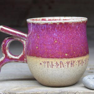 May include: A handcrafted ceramic mug with a distinctive handle. The mug has a magenta glaze on the upper half and a speckled beige base. The handle and rim have a white and magenta design. The mug has a stone next to it.