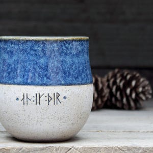 May include: A white ceramic mug with a blue glaze on the top half. The mug has a speckled finish and features a row of runes on the bottom half.
