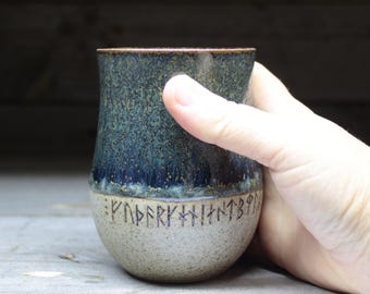 May include: A ceramic mug with a blue and green glaze and a band of black runes around the bottom. The mug is being held in a hand.
