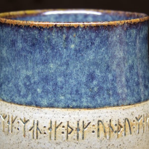 Puede incluir: Una taza de cerámica con un esmalte azul y blanco. La mitad inferior de la taza es blanca con una banda marrón alrededor de la parte superior. La mitad superior de la taza es un esmalte azul moteado con una banda marrón alrededor de la parte inferior. La taza tiene una banda de runas alrededor de la parte inferior.