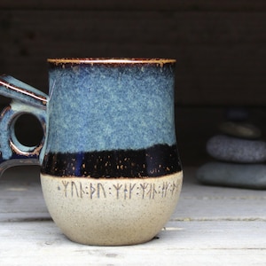 May include: A blue and brown ceramic mug with a unique handle and a band of runes around the bottom.