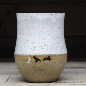 Könnte beinhalten: Eine weiße Keramiktasse mit brauner, gesprenkelter Glasur. Die Tasse hat einen Halbmond und zwei Vögel in schwarzen Silhouetten auf der unteren Hälfte.