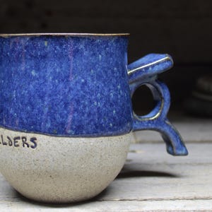 May include: A handcrafted ceramic mug, featuring a blue speckled glaze on the upper section and a beige speckled base. The word "SHOULDERS" is inscribed in black. The handle has a distinctive, artistic design.