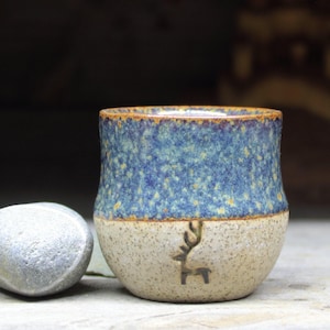 Puede incluir: Una taza de cerámica pequeña hecha a mano con un esmalte moteado azul y marrón. La taza tiene una base blanca con una silueta de ciervo marrón.