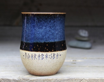 May include: A blue and white ceramic mug with black speckles and a band of black glaze. The mug has a white base with runes inscribed in black.