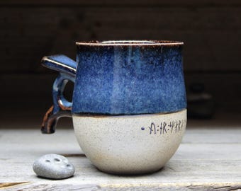 May include: A handmade ceramic mug with a blue and white glaze. The top half is a deep blue, transitioning to white at the base. The handle is a unique, angular design. The mug has a rustic, artisanal feel, perfect for coffee or tea.