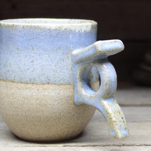 May include: A handmade ceramic mug with a unique handle design. The mug features a two-tone color scheme, with a light blue upper half and a tan lower half. The handle is also light blue and has a distinctive, angular shape. The mug is likely used for drinking beverages.