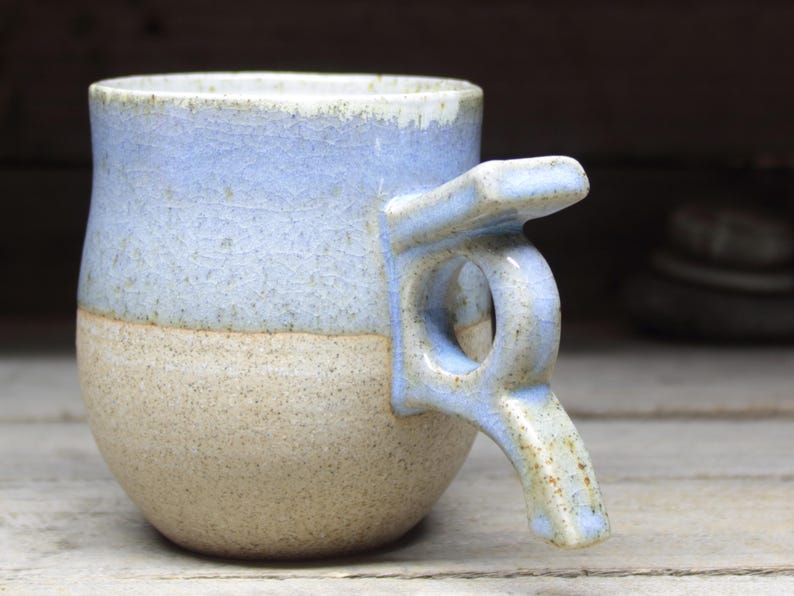 May include: A handmade ceramic mug with a unique handle design. The mug features a two-tone color scheme, with a light blue upper half and a tan lower half. The handle is also light blue and has a distinctive, angular shape. The mug is likely used for drinking beverages.