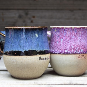 Pode incluir: Duas canecas de cerâmica com um esmalte salpicado. A caneca à esquerda é azul com uma borda preta e as palavras "Husband" escritas no lado. A caneca à direita é roxa com uma borda preta e as palavras "Wife" escritas no lado.