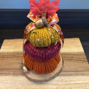 May include: A decorative stack of three knitted pumpkins in autumnal colours: orange, burgundy, and yellow. It is adorned with a red leaf, ribbon, and gold acorns. The pumpkins are displayed on a wooden slice and board, perfect for autumn.