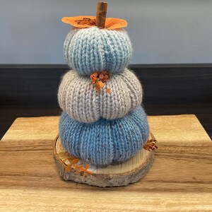 May include: A decorative stack of three knitted pumpkins in shades of blue and grey, topped with a cinnamon stick and dried orange accents. The pumpkins sit on a wooden slice, displayed on a wooden surface.