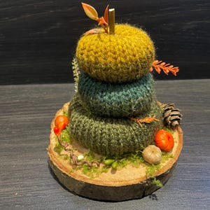 May include: A knitted autumn display with three stacked circles in yellow, green, and olive. It features miniature pumpkins, a pine cone, and foliage, set on a wooden base. A seasonal, handcrafted decoration.