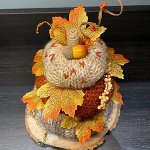 May include: A knitted pumpkin decoration with autumnal leaves and berries. The pumpkin is in shades of brown and orange, set on a wooden base. The leaves are yellow and orange. A seasonal decoration for autumn. The pumpkin is approximately 15 cm in height.