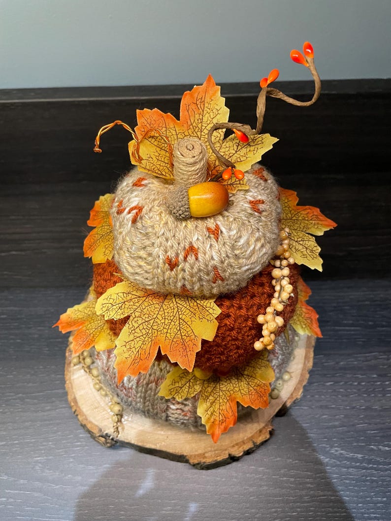 May include: A knitted pumpkin decoration with autumnal leaves and berries. The pumpkin is in shades of brown and orange, set on a wooden base. The leaves are yellow and orange. A seasonal decoration for autumn. The pumpkin is approximately 15 cm in height.