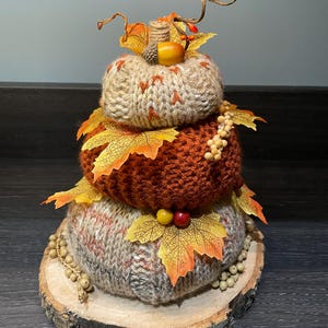 May include: A three-tiered decorative pumpkin made of knitted yarn in shades of beige, orange, and brown, adorned with fall leaves, berries, and an acorn. The pumpkin sits on a wooden slice base, perfect for autumn decor.