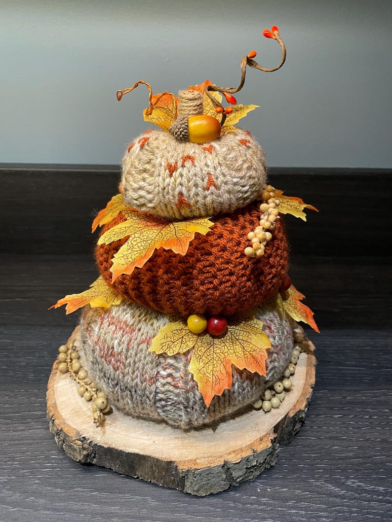 May include: A three-tiered decorative pumpkin made of knitted yarn in shades of beige, orange, and brown, adorned with fall leaves, berries, and an acorn. The pumpkin sits on a wooden slice base, perfect for autumn decor.