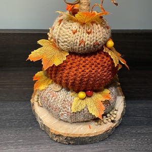May include: A decorative stacked pumpkin, featuring three knitted pumpkins in beige and orange hues, decorated with artificial autumn leaves and berries. The pumpkins rest on a wooden slice base, evoking a rustic autumnal feel.