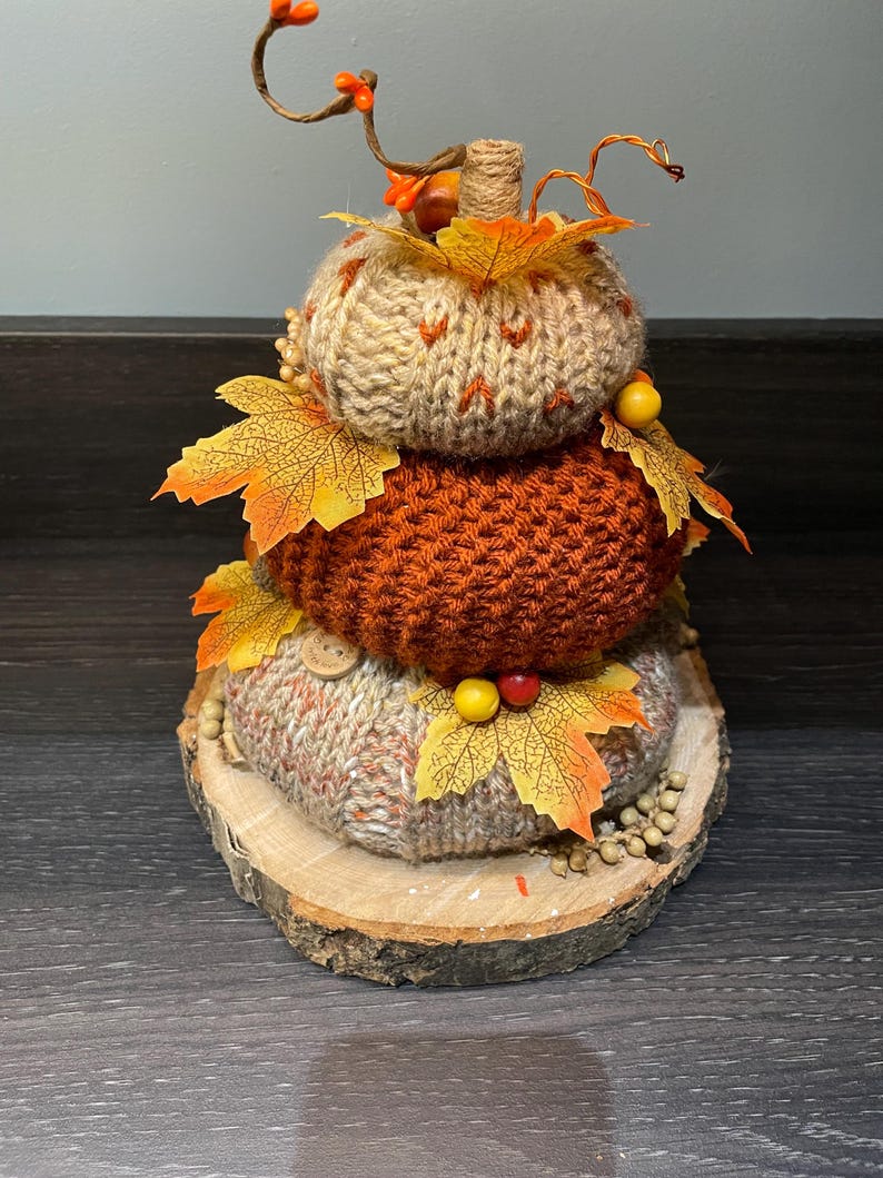 May include: A decorative stacked pumpkin, featuring three knitted pumpkins in beige and orange hues, decorated with artificial autumn leaves and berries. The pumpkins rest on a wooden slice base, evoking a rustic autumnal feel.