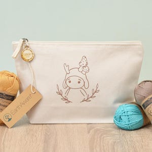 Könnte beinhalten: Eine cremefarbene Canvas-Tasche mit einer Pilz-Charakter-Illustration, einem Reißverschluss und einem Holzanhänger. Die Tasche ist von Garnknäueln in Gelb, Türkis und Taupe umgeben, mit Scheepjes Catona-Etiketten und einem EarthAware-Anhänger.