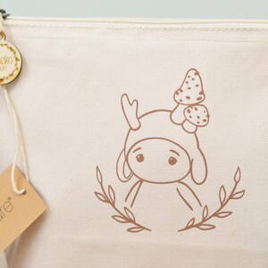 Puede incluir: Bolsa de cosm&eacute;ticos de lona color crema con una ilustraci&oacute;n marr&oacute;n de un personaje de dibujos animados con un sombrero de seta. La bolsa tiene cierre de cremallera y una etiqueta de madera que dice "Sameko Design". Tambi&eacute;n hay una etiqueta marr&oacute;n que dice "hAware".