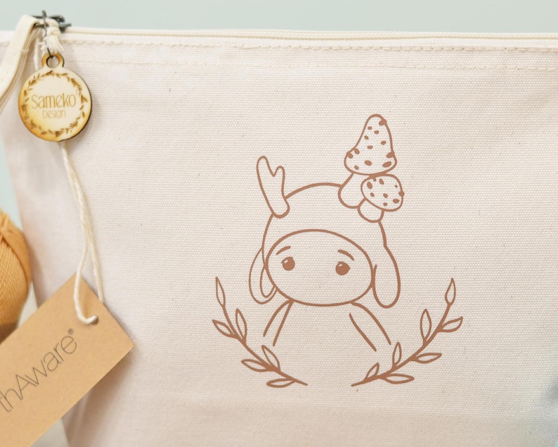 Puede incluir: Bolsa de cosm&eacute;ticos de lona color crema con una ilustraci&oacute;n marr&oacute;n de un personaje de dibujos animados con un sombrero de seta. La bolsa tiene cierre de cremallera y una etiqueta de madera que dice "Sameko Design". Tambi&eacute;n hay una etiqueta marr&oacute;n que dice "hAware".