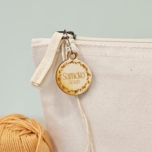 Puede incluir: Primer plano de una bolsa de lona color crema con cremallera. Una etiqueta de madera con el texto "Sameko Design" est&aacute; sujeta a la cremallera. La etiqueta es redonda y presenta un dise&ntilde;o de hojas. Una madeja de hilo amarillo en primer plano.