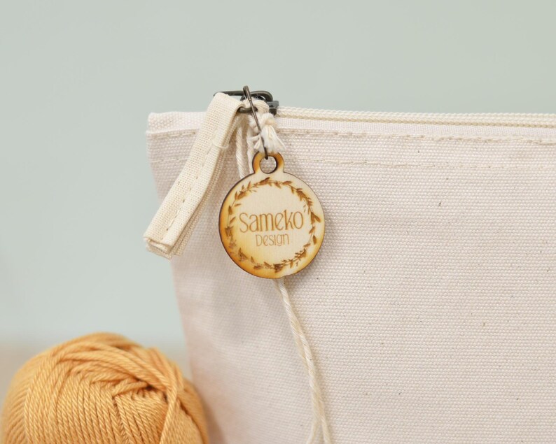 Puede incluir: Primer plano de una bolsa de lona color crema con cremallera. Una etiqueta de madera con el texto "Sameko Design" est&aacute; sujeta a la cremallera. La etiqueta es redonda y presenta un dise&ntilde;o de hojas. Una madeja de hilo amarillo en primer plano.