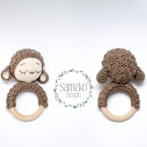Pode incluir: Um chocalho de cordeiro de crochê marrom com um anel de madeira. O cordeiro tem um rosto branco com olhos e boca bordados.