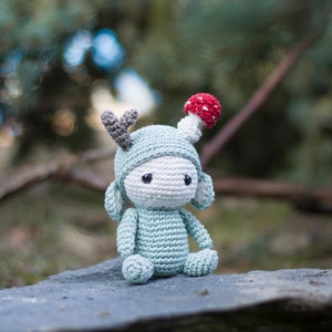 May include: A crocheted doll in a seated position, featuring a light blue body and a white face. The doll wears a light blue hat with gray antlers and a red and white mushroom on top. The doll has black eyes and is made of yarn.