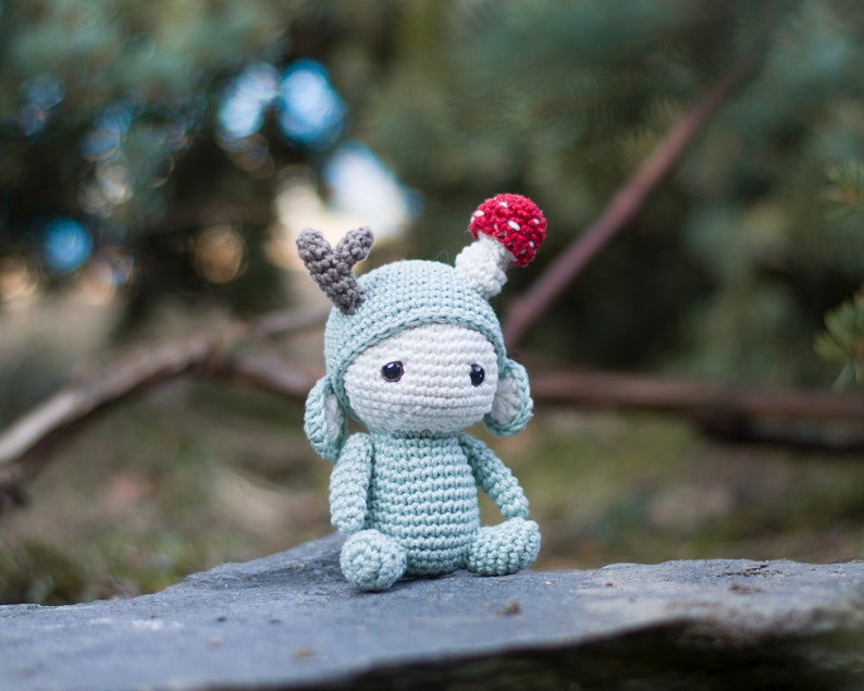 May include: A crocheted doll in a seated position, featuring a light blue body and a white face. The doll wears a light blue hat with gray antlers and a red and white mushroom on top. The doll has black eyes and is made of yarn.