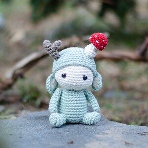 May include: A handmade crochet doll in a light blue outfit with a matching hat and gray antlers. The doll has a white face and a red and white mushroom on top of the hat. The doll is sitting on a gray stone.